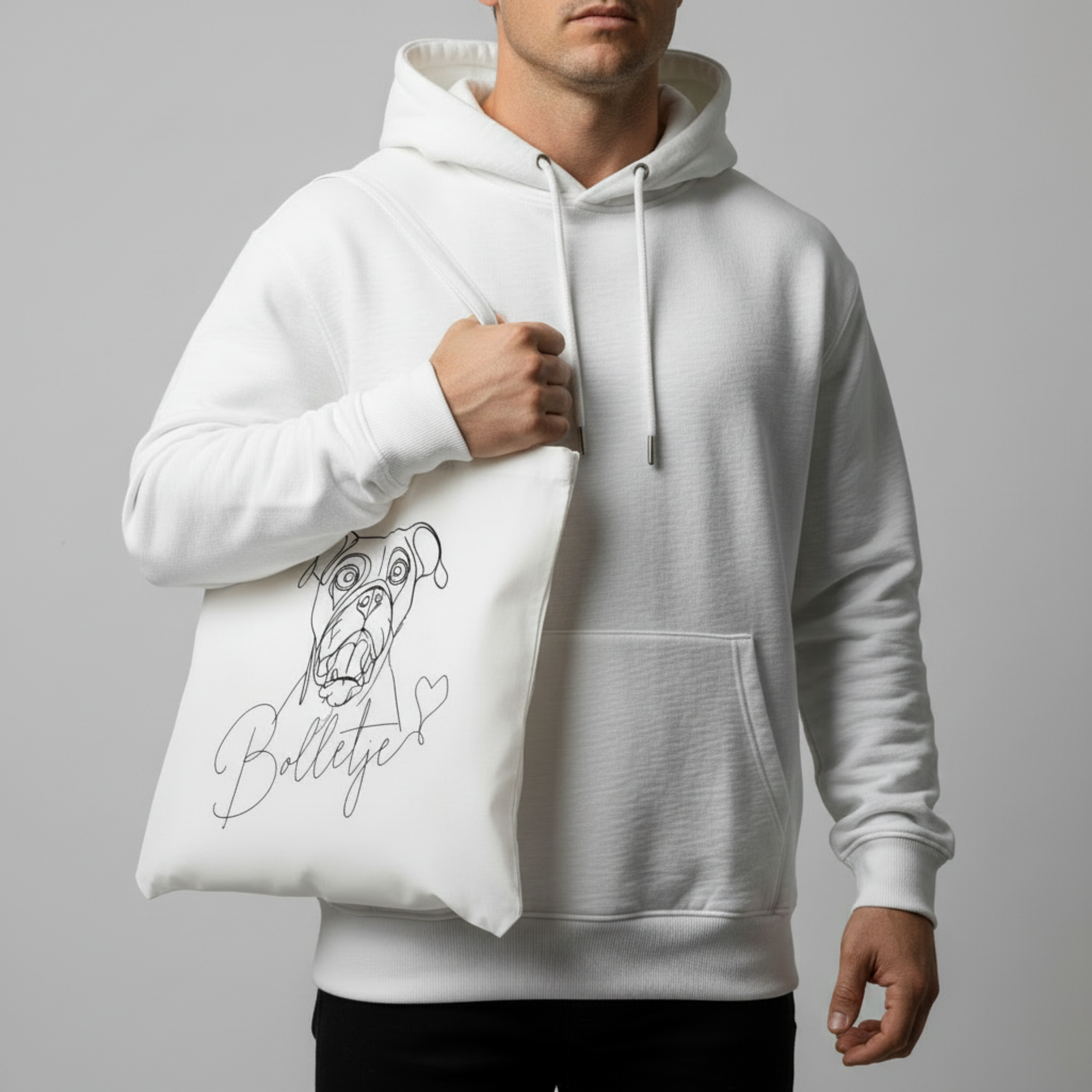 Person wearing a white hoodie holding a tote bag with a dog illustration and text. Just Groom Me – Trimsalon Alphen aan den Rijn | Professional dog grooming & personalized pet apparel.