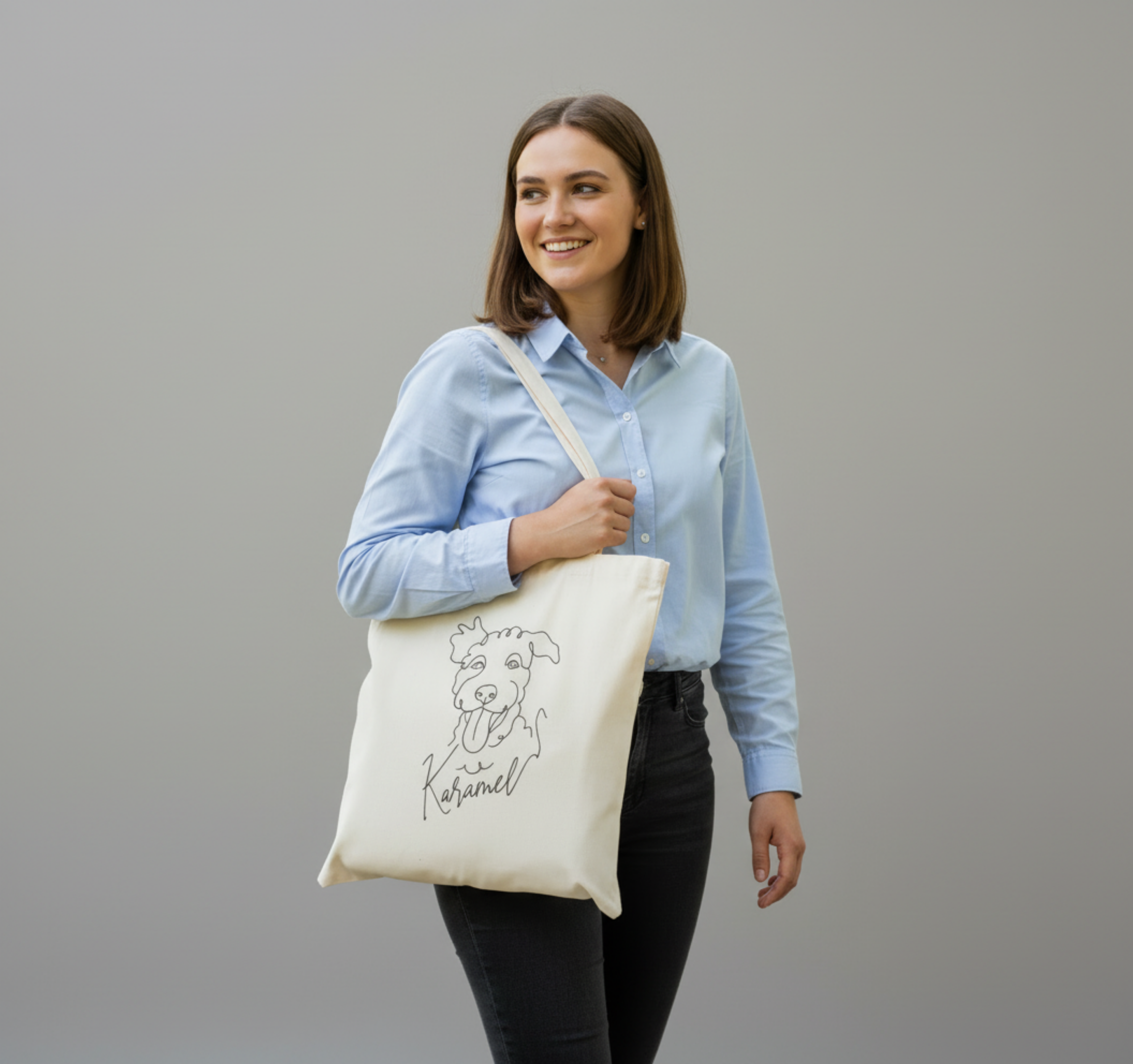 Woman holding a tote bag with a dog illustration and text against a gray background. Just Groom Me – Trimsalon Alphen aan den Rijn | Professional dog grooming & personalized pet apparel.